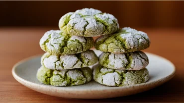 Stack of vibrant green matcha crinkle cookies dusted with powdered sugar on a white ceramic plate