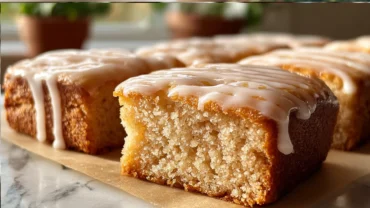 Close-up of fluffy baked maple donut bars topped with a shiny maple glaze, arranged on a cooling rack in a bright kitchen, with a drizzle of icing and a jar of maple syrup in the background.