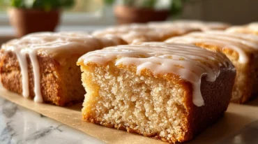 Close-up of maple donut bars topped with a shiny maple glaze, arranged on a cooling rack in a bright kitchen, with a drizzle of icing and a jar of maple syrup in the background.