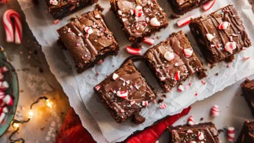 Close-up of Fudgy Peppermint Brownies topped with crushed candy canes and white chocolate drizzle on parchment paper.