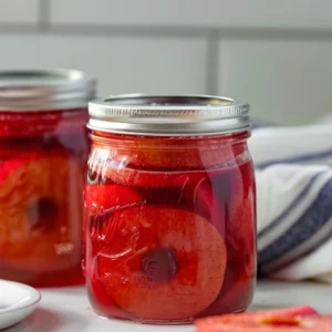 Two glass jars filled with glossy red spiced apple rings in syrup, styled on a white wooden table with a folded white-and-blue kitchen towel and sliced apples in front — cozy homemade fall treat with cinnamon and clove.