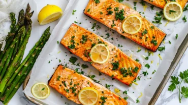 Top-down view of baked salmon fillets with lemon slices and roasted asparagus on a parchment-lined tray.