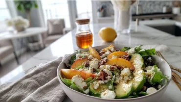 Close-up of a peach avocado salad in a white ceramic bowl with mixed greens, sliced peaches, avocado, feta cheese, and toasted pecans, drizzled with honey-lemon dressing under soft natural daylight on a marble countertop.