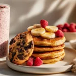 Close-up photo of chocolate chip cookies stacked, fluffy banana pancakes topped with maple syrup and banana slices, and a berry smoothie with raspberries and chia seeds, styled on a white marble countertop in bright modern natural light.