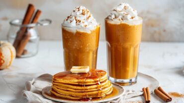 Two pumpkin smoothies topped with whipped cream beside a stack of pumpkin pancakes with syrup on a modern white background clean, cozy fall breakfast.