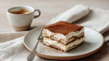 Slice of tiramisu without raw eggs on ceramic plate with espresso cup beside it